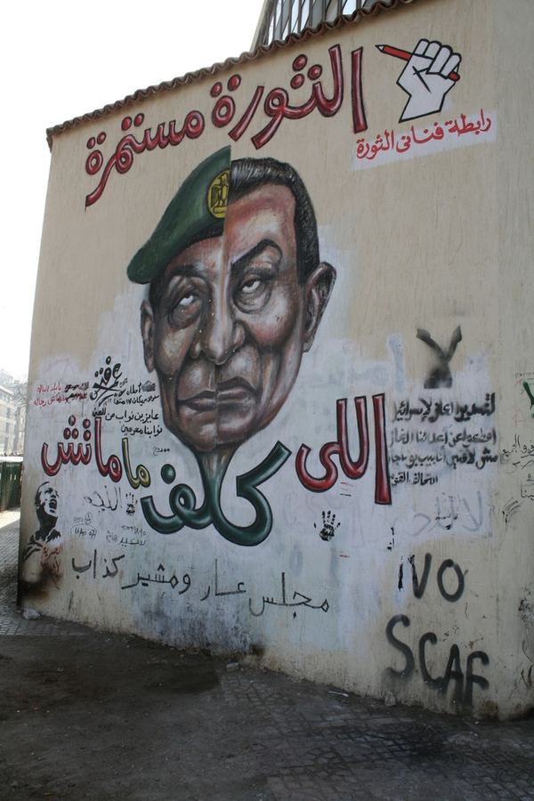 A mural of a divided military portrait beside Arabic revolutionary slogans