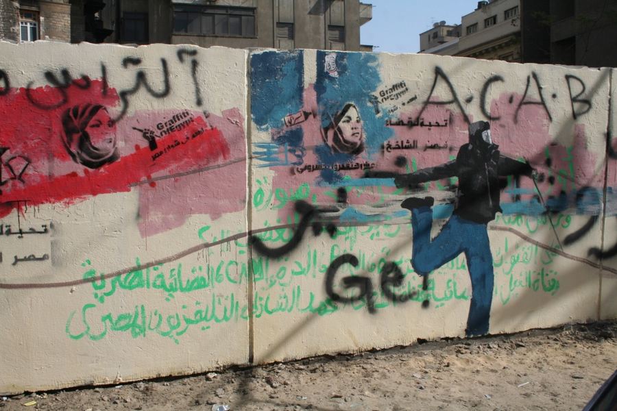 A concrete barrier covered in Arabic graffiti, stencilled faces and the letters ACAB