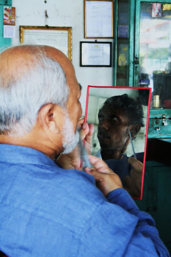 A barber shaving a customer, both reflected in a red-framed mirror