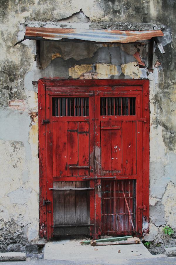 A weathered red timber door in a crumbling shophouse facade