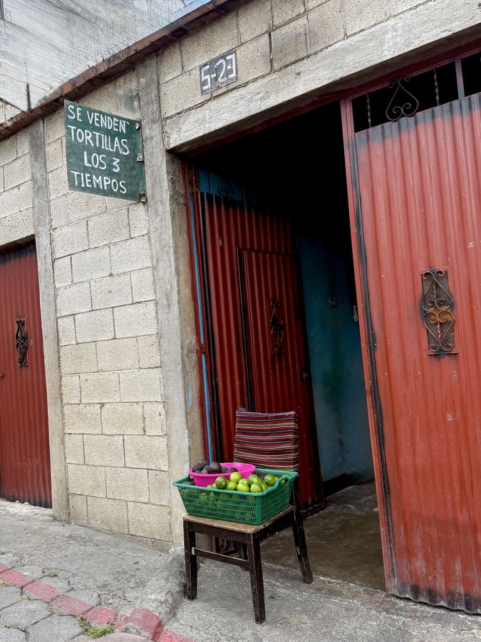 A hand-painted tortilla sign beside red doors and a crate of produce