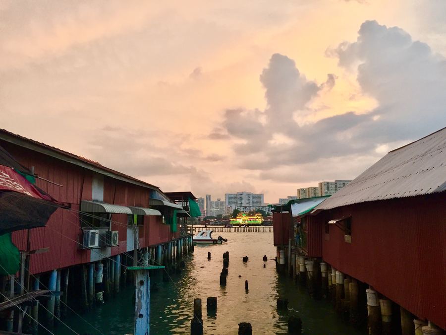 Stilt houses lining a channel at sunset, with a city skyline beyond