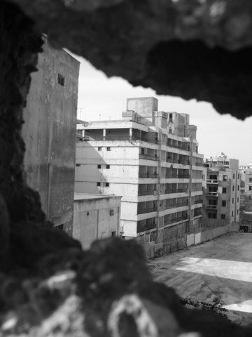 Apartment blocks framed through a jagged gap in a wall, in black and white